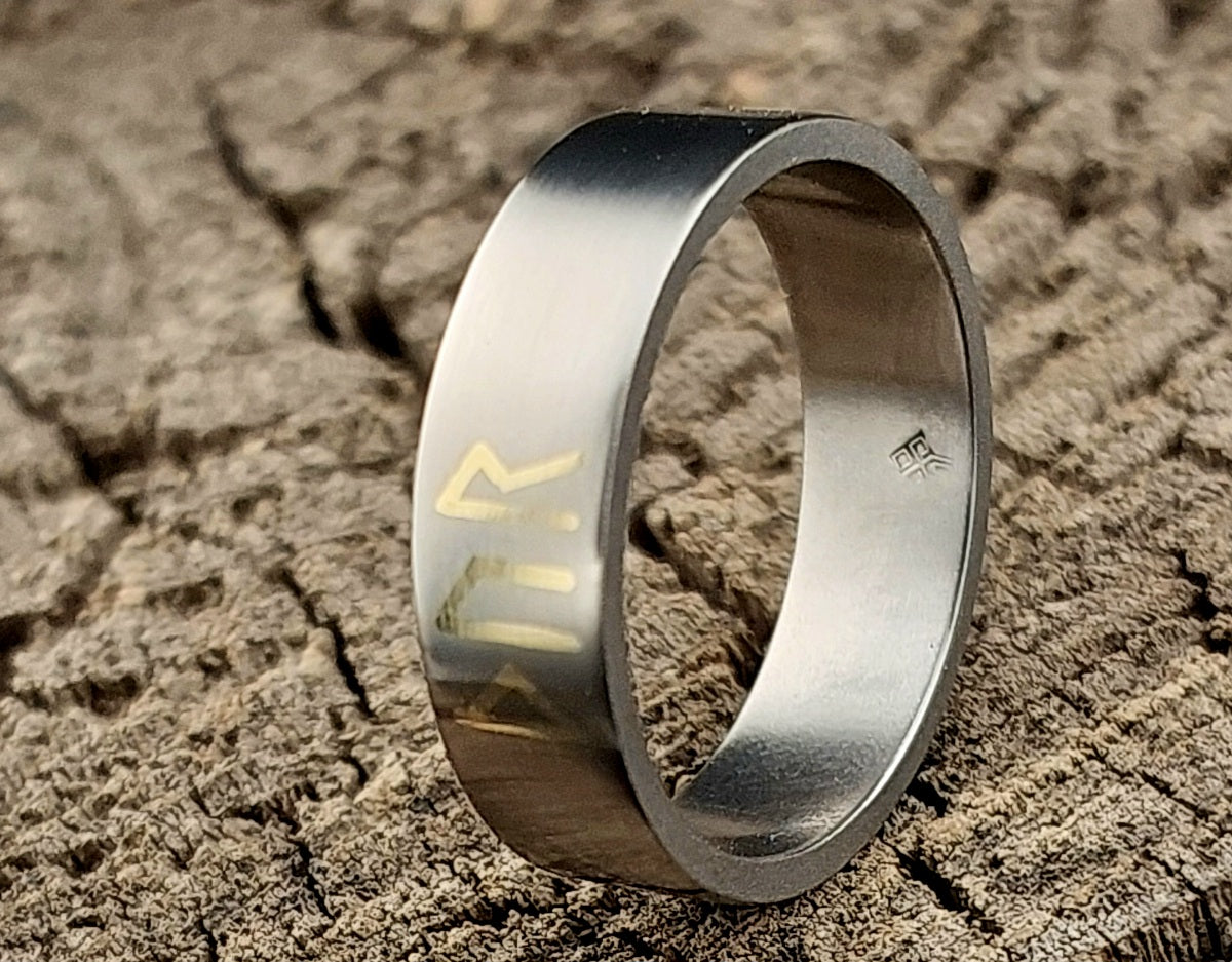 Close-up of the satin brushed titanium surface with polished silver or brass rune inlay reflecting the light.